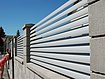 Vue rapprochée d'une clôture de jardin à lamelles couleur gris aluminium sur socles muraux, avec une jeune fille qui ouvre le portillon