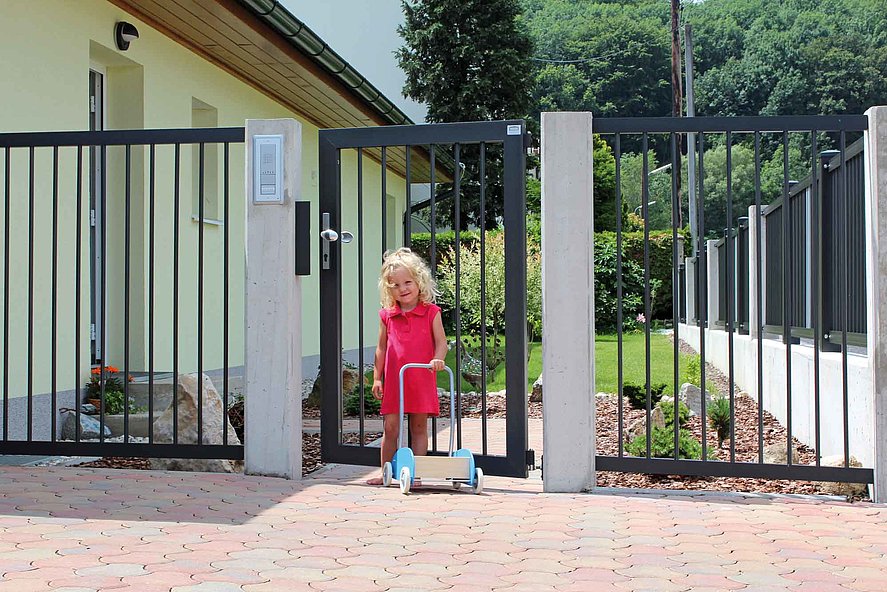 Jeune fille sur l'entrée à pavés, devant un portillon de jardin couleur anthracite assorti à la clôture à barres