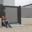 Jeune garçon adossé sur le portillon de jardin à lamelles grises en aluminium sur socle mural blanc