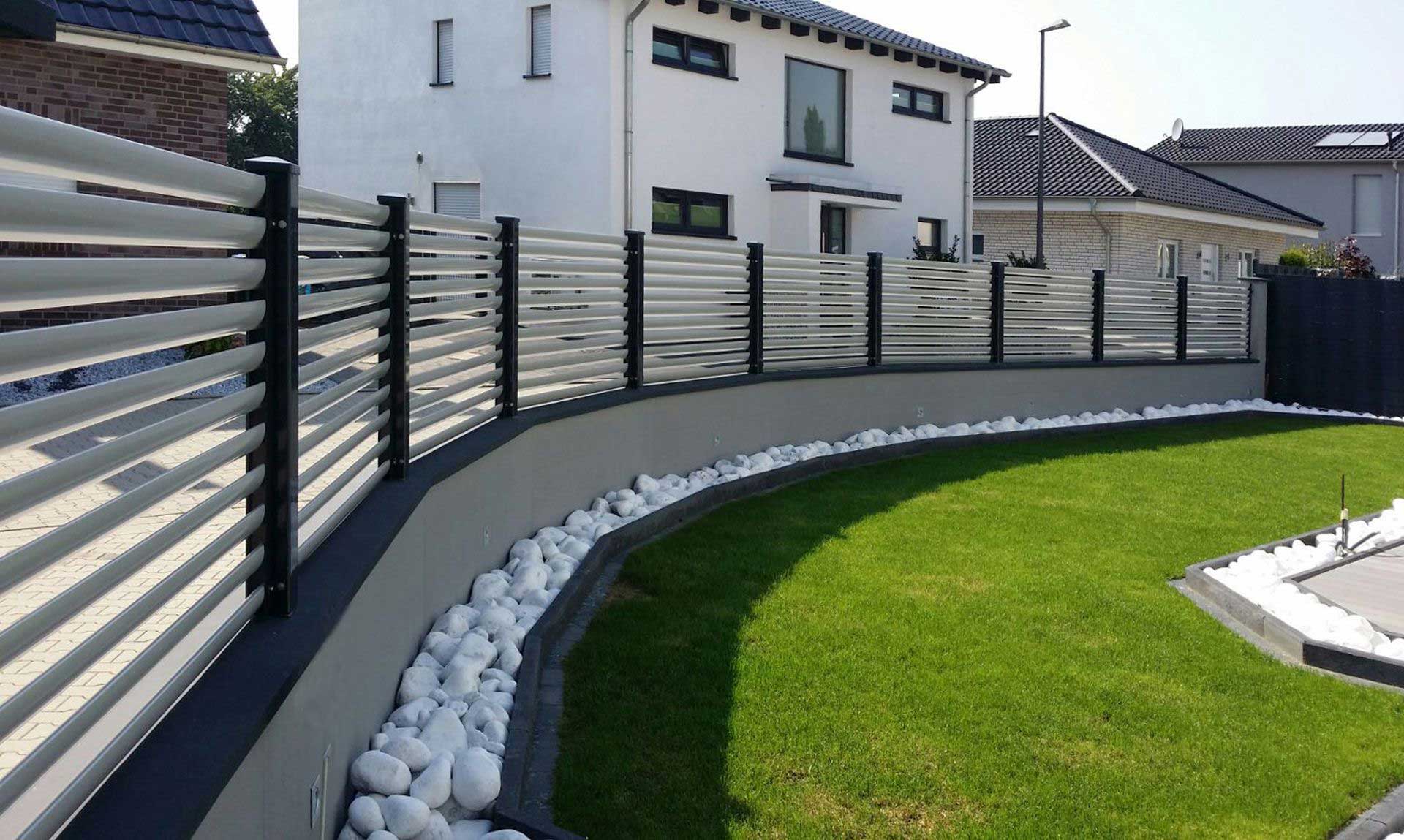 Clôture en blanc sur toute l'image, devant la clôture une grande pelouse dans un jardin, derrière la clôture une grande maison blanche